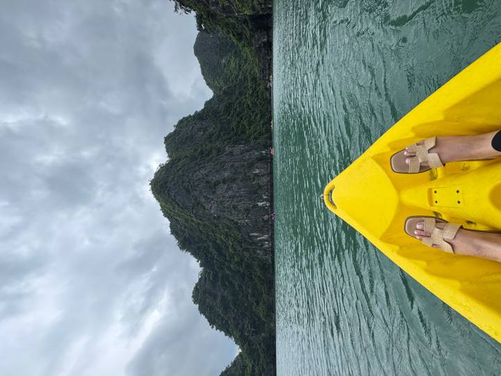 View from a yellow kayak bow with a traveller’s feet pointing toward emerald waters and towering limestone cliffs.