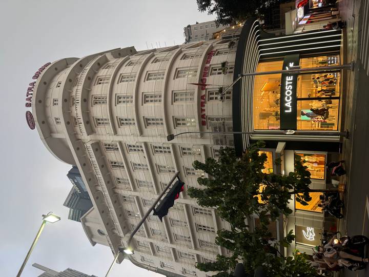Tall white SATRA Group building with a glowing Lacoste store at street level during dusk in a busy city.