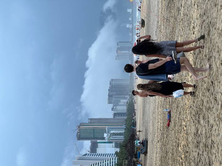 Sandy beach with travellers walking while high-rise buildings of Da Nang rise under a dramatic cloudy sky.