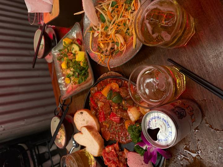 Close-up of Vietnamese dishes: sizzling stew, fresh salads, bread, beer and iced drinks on a rustic table.