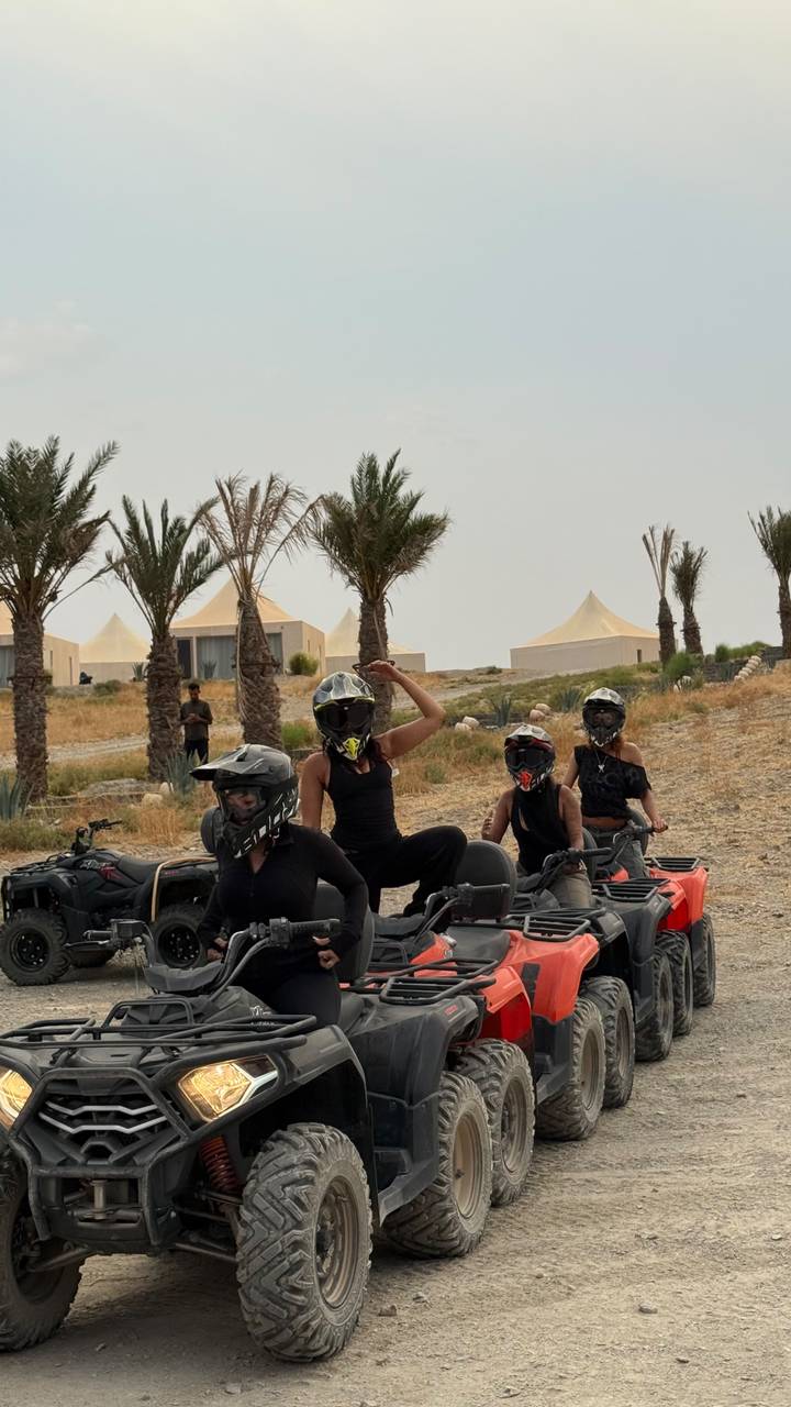 Helmeted riders on quad bikes strike playful poses in front of desert glamping tents and palm trees.