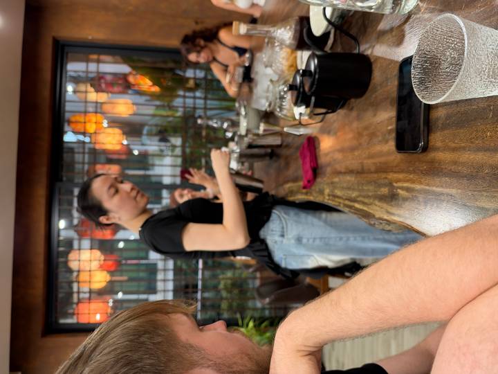 Barista explains brewing techniques behind a cafe counter with patrons watching
