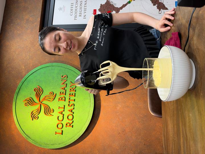 Woman whisking creamy egg coffee under a cafe sign that reads 'Local Beans Roastery'