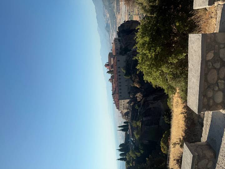 Distant view of a Meteora cliff-top monastery framed by trees and hazy valley below.