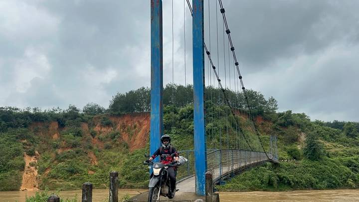 Motorbike rider on dirt bike crossing narrow blue suspension bridge over muddy river