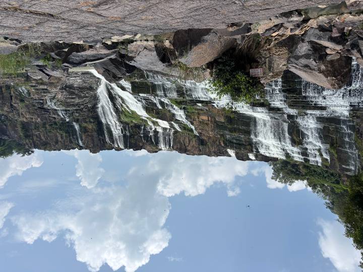Wide multi-tiered waterfall cascading over dark rock formations under blue sky with clouds