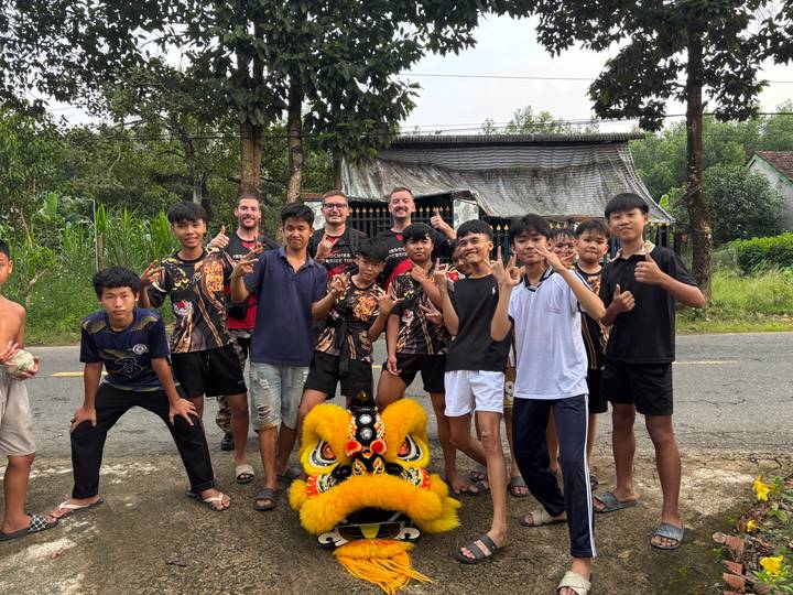 Large group of local children and travellers posing playfully on roadside next to lion dance head
