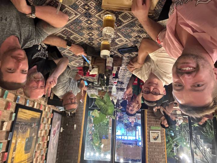 Travellers gathered around restaurant table enjoying drinks and meal, raising hands for photo