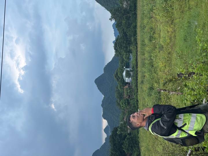 Motorbiker standing in a field smoking, with green peaks and cloudy sky in the background