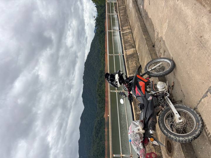Off-road motorbike parked on a concrete dam overlooking a calm reservoir and forested hills under grey skies