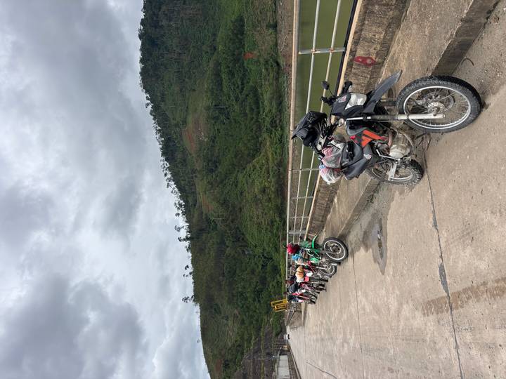 Row of dirt bikes parked on a concrete bridge beside a forested reservoir bank under overcast skies