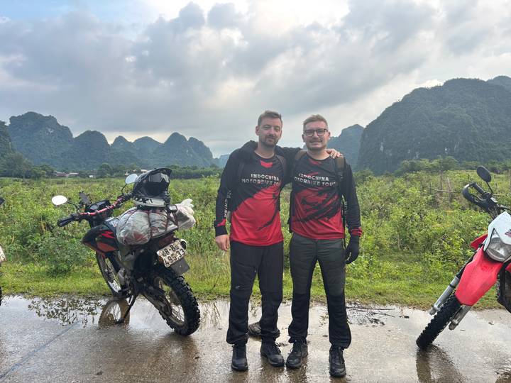 Two motorcyclists in matching jerseys pose with their bikes against limestone karst mountains after rain