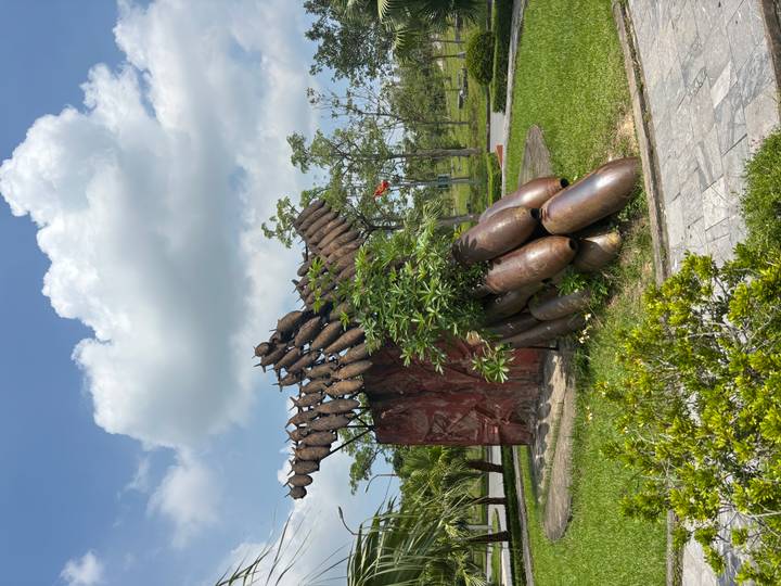 Outdoor memorial sculpture made of stacked aerial bombs surrounded by manicured grass and blue sky