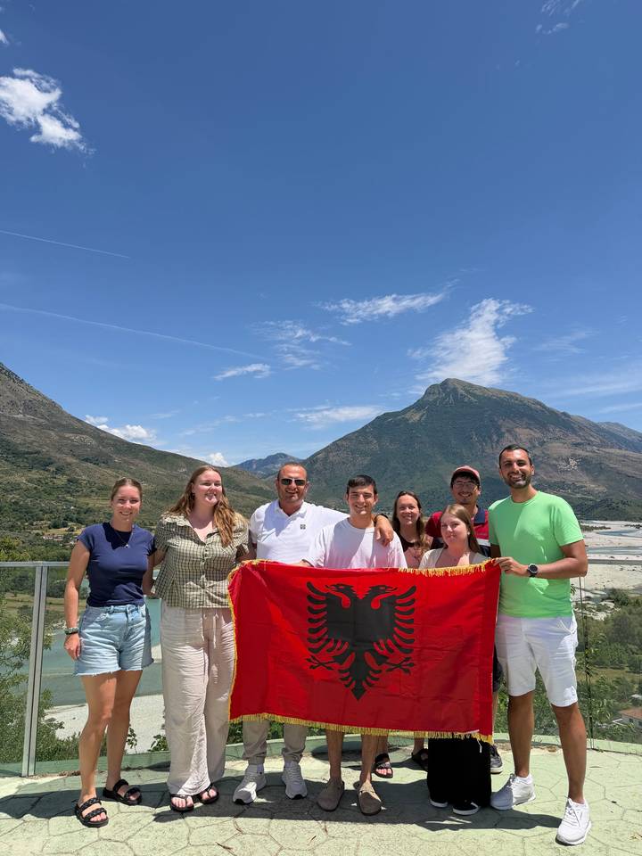 Travellers hold an Albanian flag against a dramatic mountain backdrop under a bright blue sky.