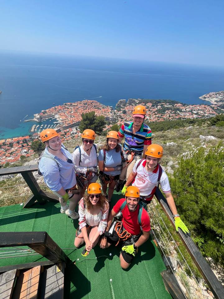 Thrilled travellers in helmets stand on a mountaintop zipline platform with aerial view of Dubrovnik’s old town and sea below.