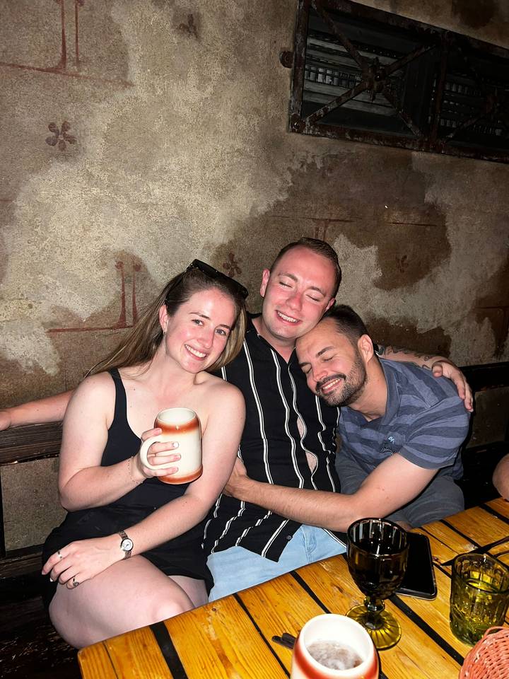 Three friends sit closely in a cozy bar, smiling and holding a mug.