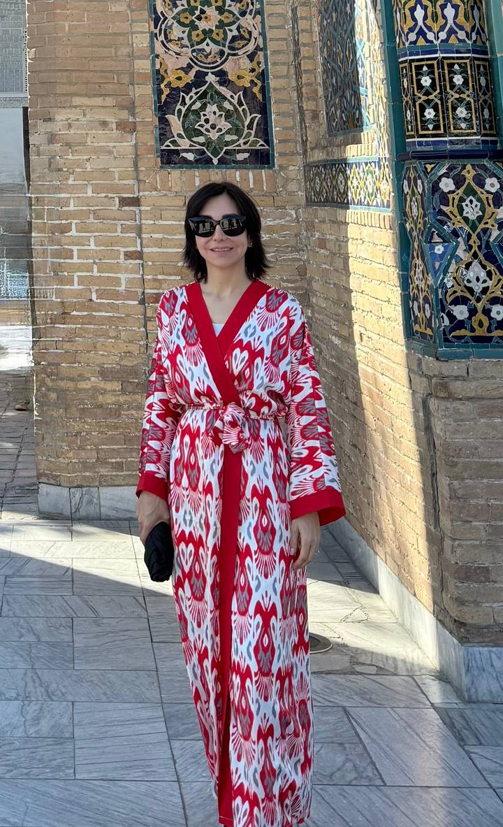 Young woman wearing a vibrant red-and-white ikat robe poses beside an ornate brick wall with tiled details.