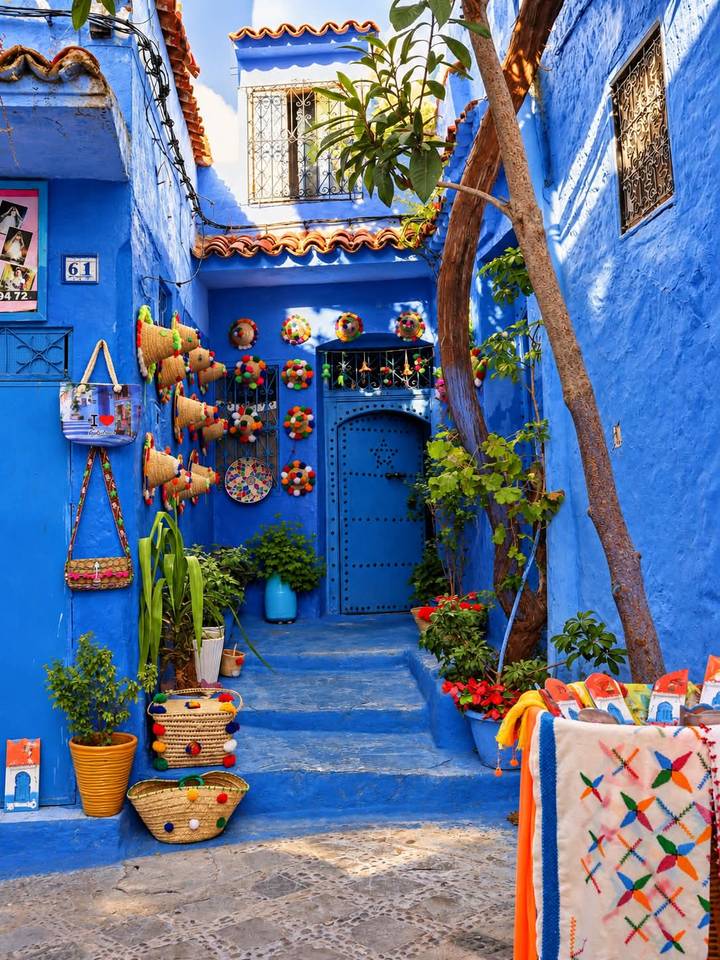 Vibrant blue alley in Chefchaouen decorated with colourful hats, plants and a patterned door.