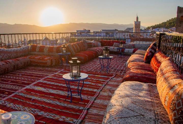 Rooftop terrace with colourful Berber carpets and cushions overlooking a minaret at sunset.