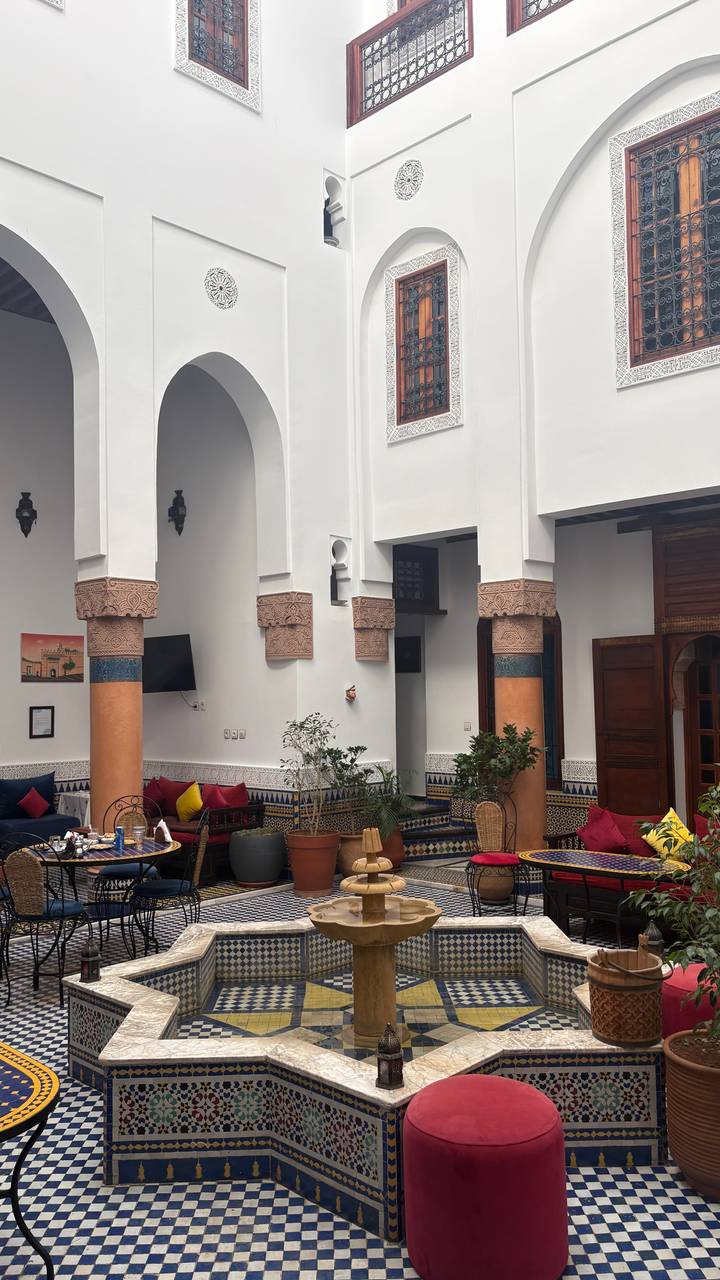 Traditional riad courtyard with carved columns, tiled walls and colourful cushions around a fountain.