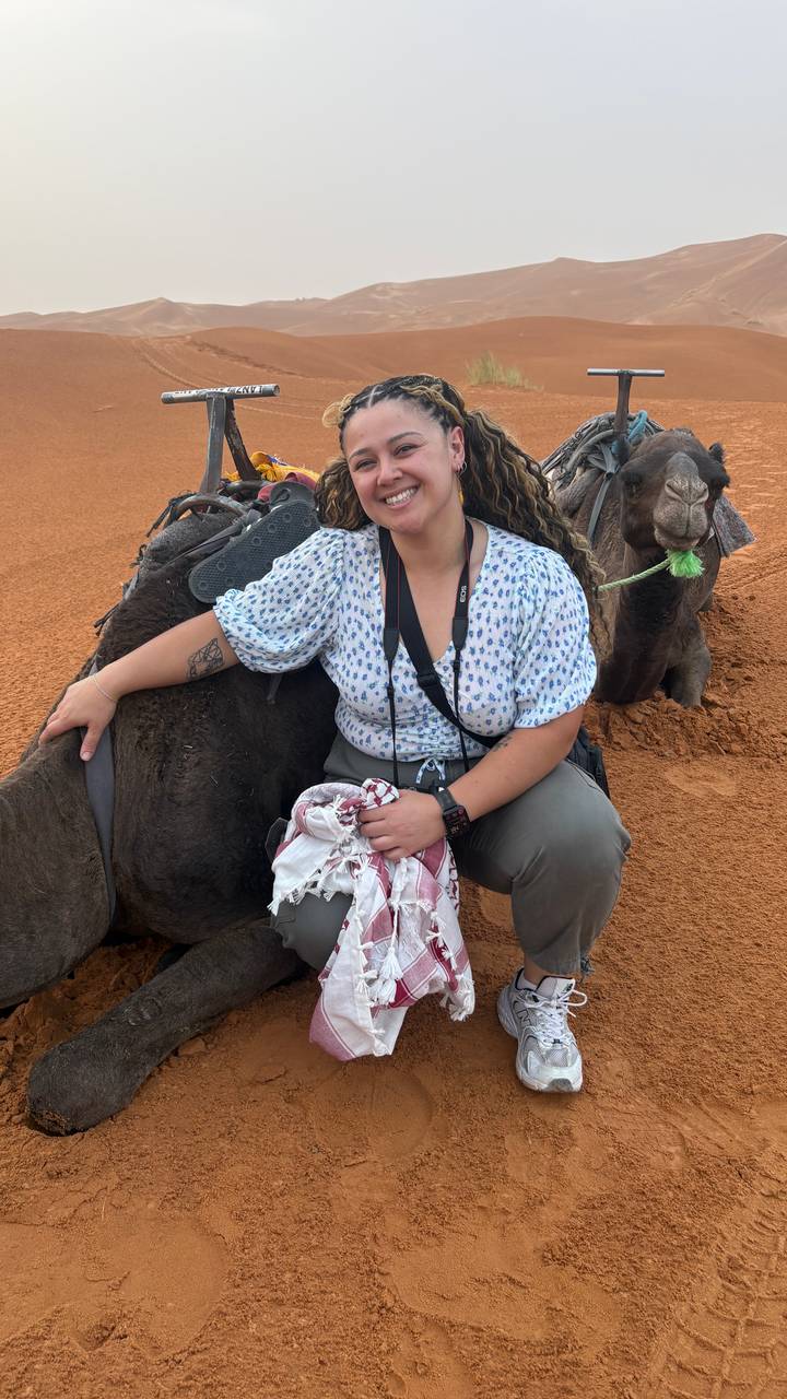Smiling traveller sits side-saddle on a resting camel in the orange desert sand.