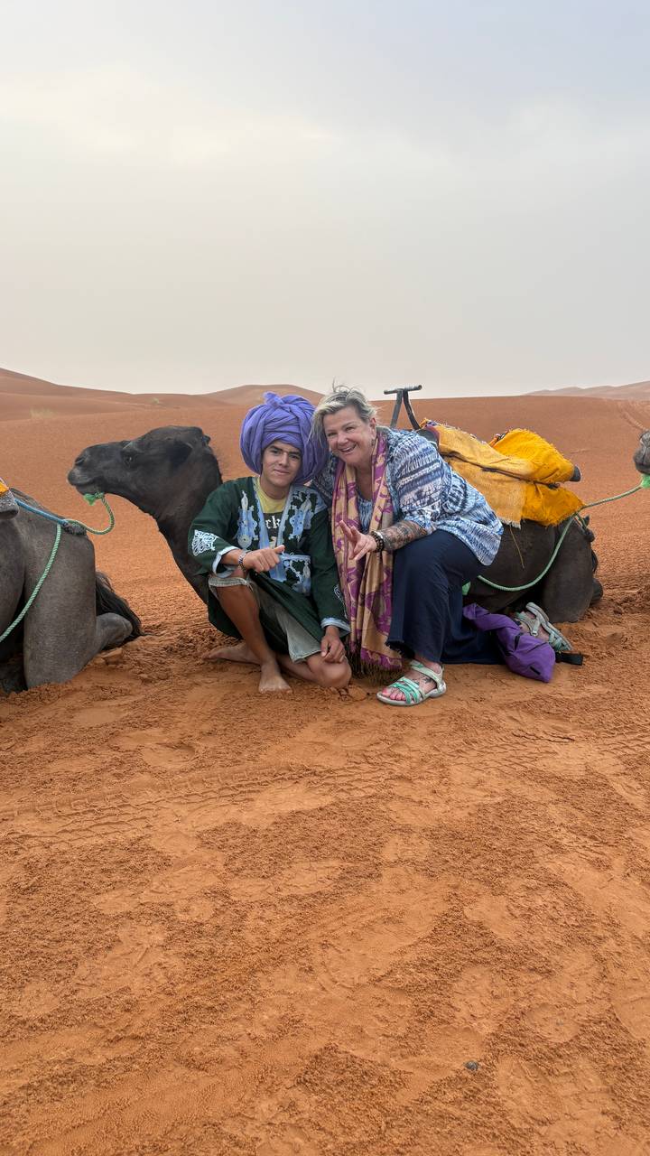 A young local man in traditional attire and an older female tourist kneel beside resting camels on red desert sand.