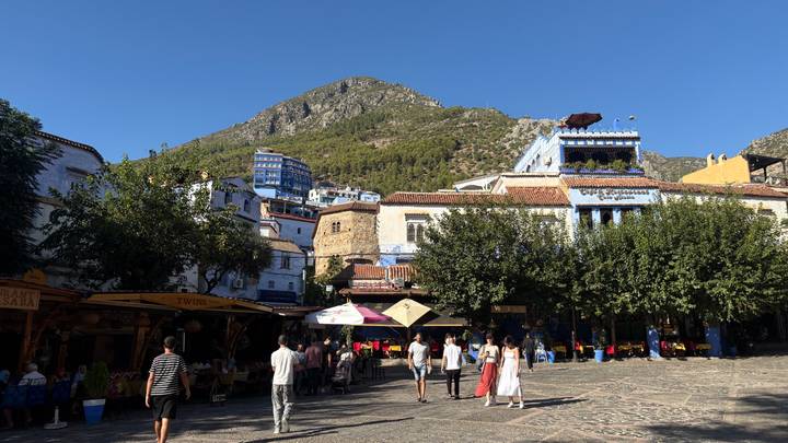 Sunny plaza framed by white-and-blue buildings with a mountain backdrop where locals and tourists stroll.