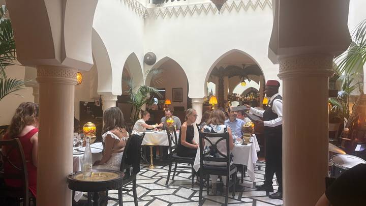 Elegant restaurant atrium with white arches where diners enjoy meals served by a waiter in traditional vest and fez.