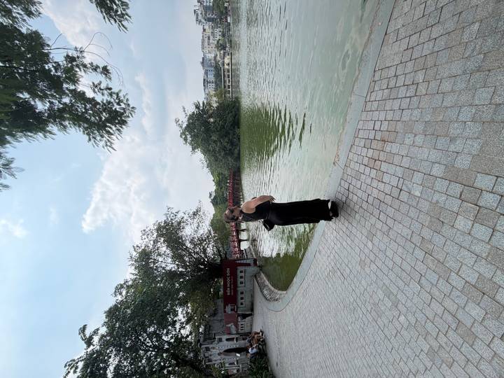 Tourist in black dress gazes across Hoan Kiem Lake toward the iconic red bridge.