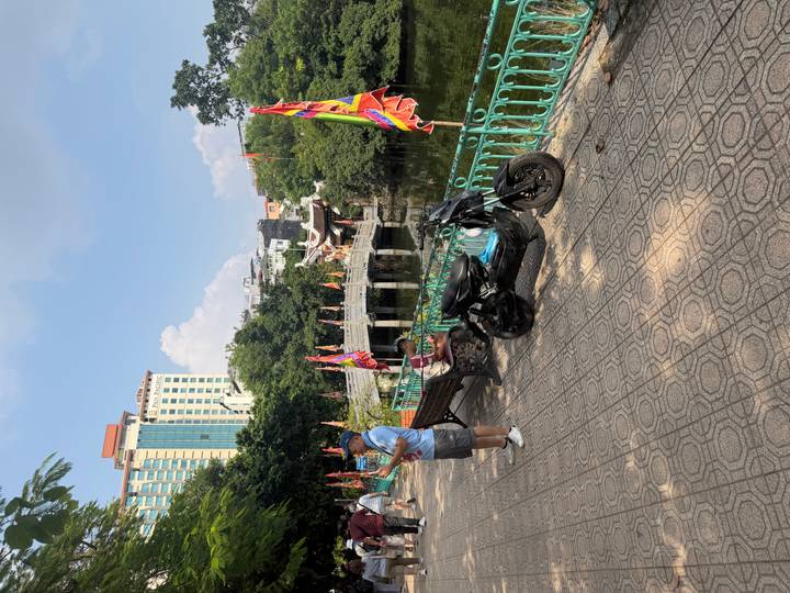 Urban park scene with scooters, colourful flags and a stone bridge over a pond under bright sun.