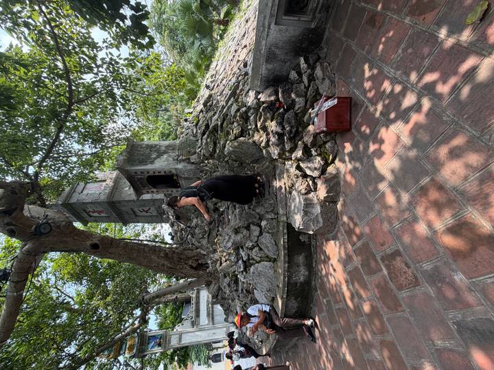 Visitor climbs rocky mound toward a small stone shrine shaded by large trees.
