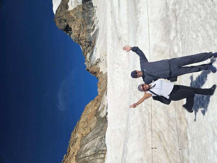 Happy travellers raising arms on a sun-lit glacier with rugged peaks under a deep blue sky.