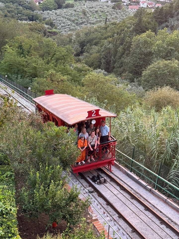 Open-air funicular car carrying tourists up a green hillside.
