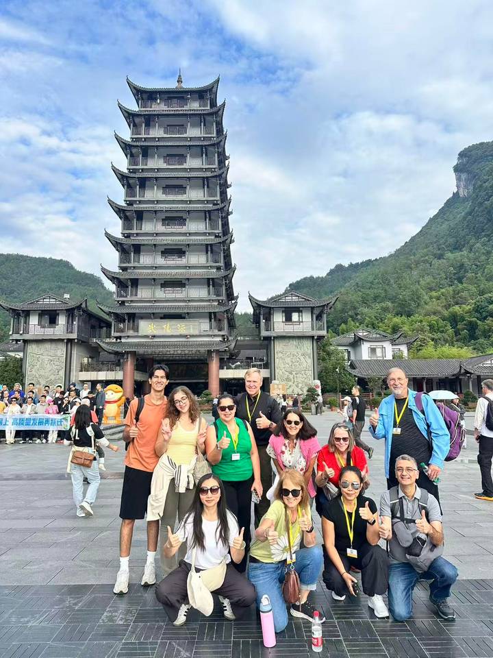 Smiling tourists stand before a multi-story pagoda gate backed by forested mountains.