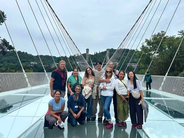 Visitors stand on a glass suspension bridge with cables stretching overhead and forested hills beyond.