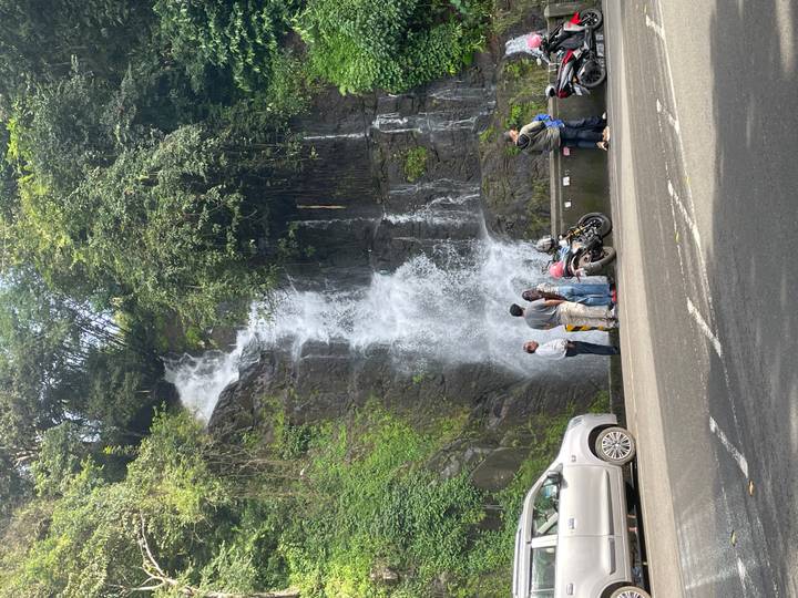 Travelers stop beside parked motorcycles to admire a roadside waterfall tumbling down a mossy cliff.