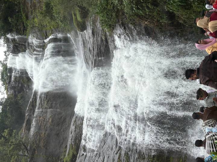 Crowds gather at the base of a tall, multi-drop waterfall plunging down a rocky cliff.