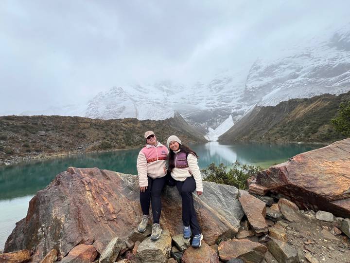 Two hikers pose on colorful rocks by a turquoise glacial lake with snowy peaks behind