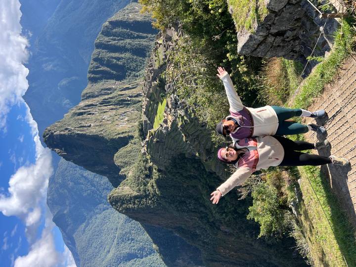 Two excited travelers raise their arms on a terrace overlooking the iconic ruins and peak of Machu Picchu.