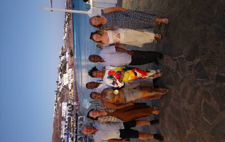 Group of friends dressed for an evening out pose beside the waterfront of Mykonos as lights twinkle in the distance.