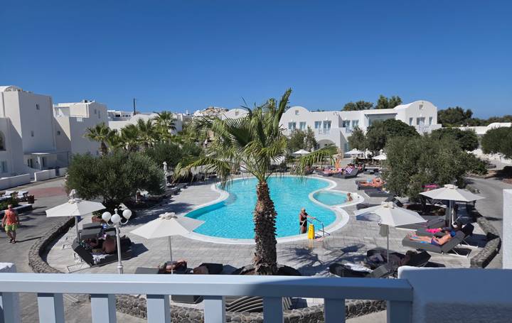 Sparkling hotel pool surrounded by white Cycladic buildings and palm trees under a bright Santorini sky.
