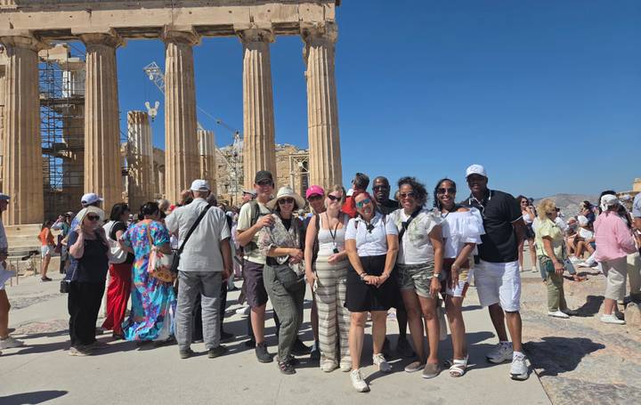 Large tour group poses in front of Acropolis columns amid busy crowds on a sunny day in Athens.