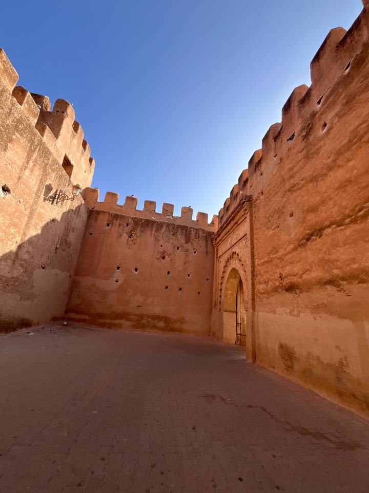 An open courtyard inside high crenellated walls, showing textured adobe surfaces lit by strong sunlight.
