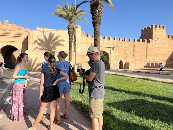 A family of travellers stands outside ancient ramparts and palm trees while a photographer captures the scene.