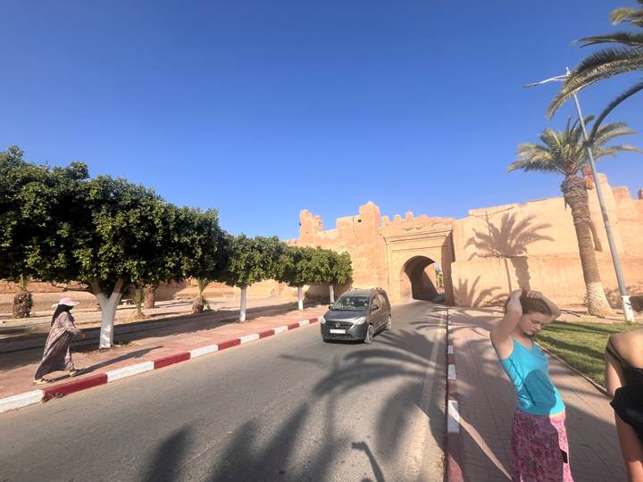 Street scene outside adobe walls with palms, a car, and casual pedestrians under bright sun.