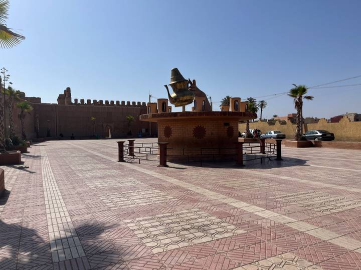 Open square with a giant teapot sculpture and fortress walls in the background on a sunny day.