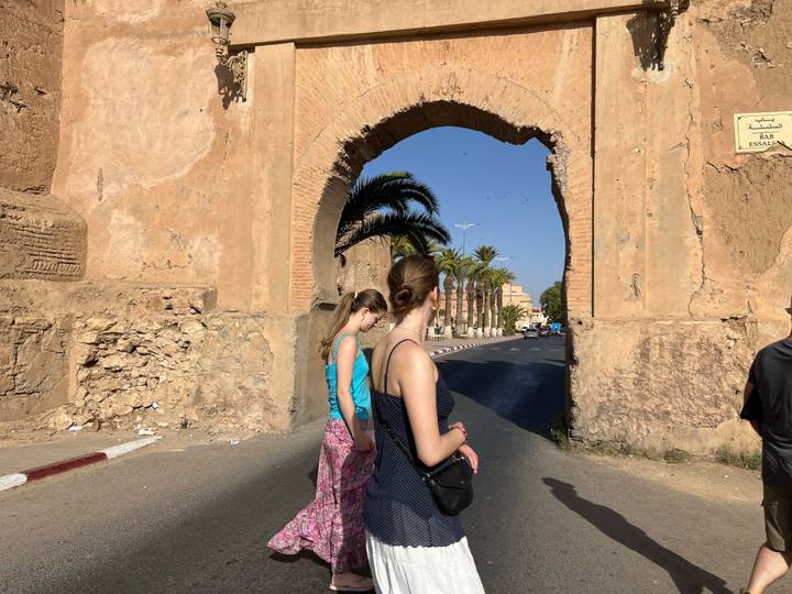 Two young travellers walk under a historic adobe arch into the old city.