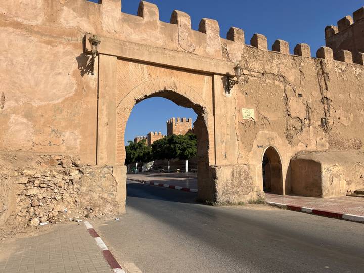 Stone archway leading through fortified walls with distant tower and clear blue sky.