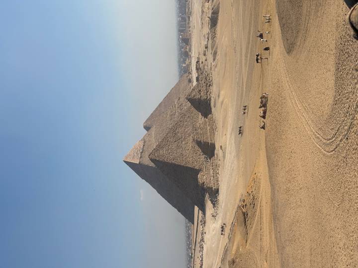 Classic view of the Pyramids of Giza rising from the desert plain with camel riders in the foreground under a clear blue sky.