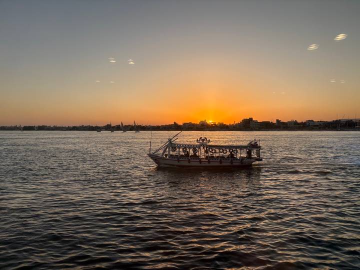 Silhouetted motorboat on the Nile at sunset with golden sky reflecting on the water and feluccas in the background.